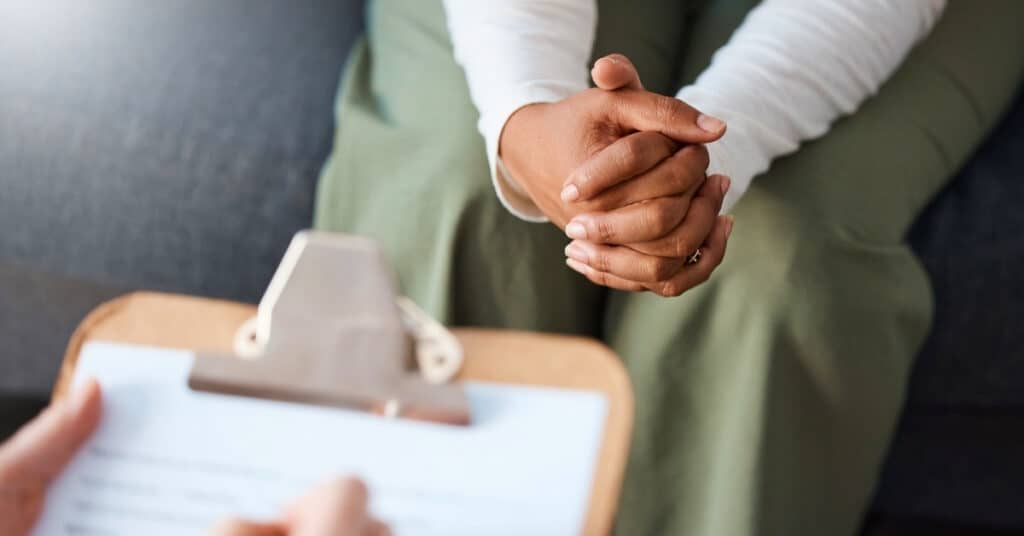 A person sits with hands clasped on their lap while another person holds a clipboard, suggesting a conversation or counseling session.