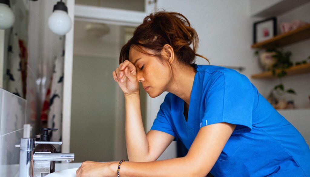 A woman in blue scrubs stands in a bathroom, leaning on the sink with her eyes closed and hand on her forehead, appearing stressed or fatigued.