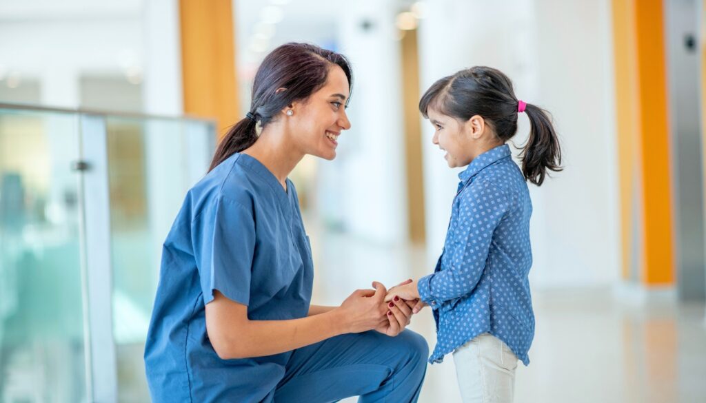 A nurse in blue scrubs kneels and smiles while holding hands with a young girl in a hospital or clinic hallway.