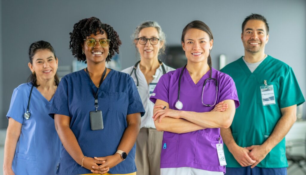 Five travel nurse in scrubs in different specializations stand together in a hospital setting, looking at the camera and smiling.