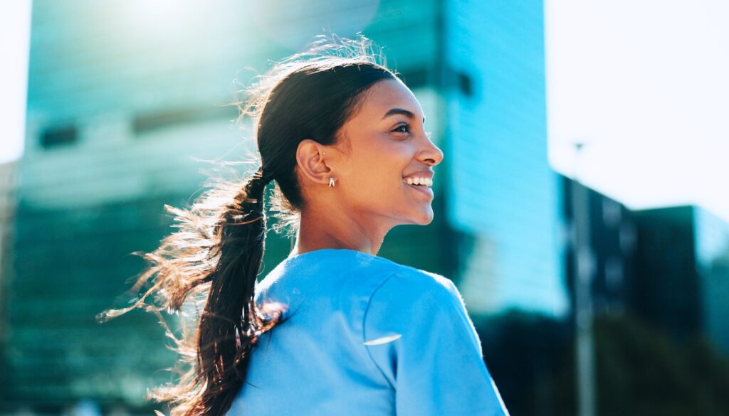 A woman in a blue shirt smiles outdoors with a modern glass building in the background and sunlight shining behind her.