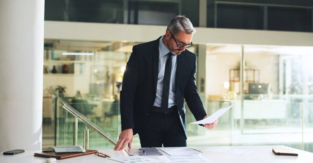 A man in a suit stands at a desk, reviewing documents in a modern office setting with large windows in the background.
