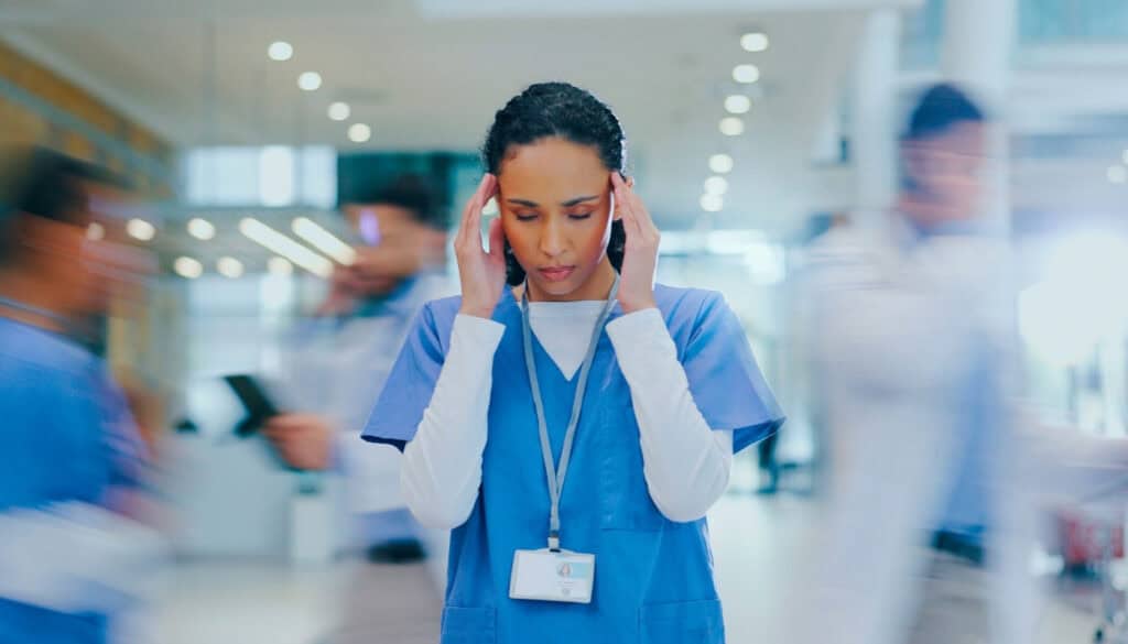 A nurse in scrubs stands with her hands on her temples, appearing stressed, while people in medical uniforms move quickly around her in a hospital corridor.