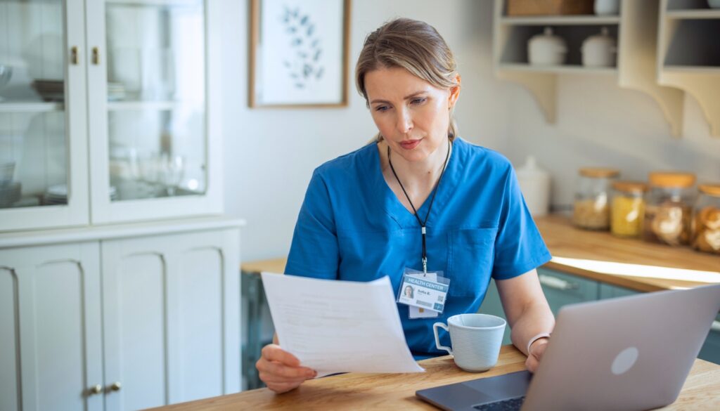 A woman in scrubs sits at a kitchen table, reading a document, with a laptop and a cup on the table.