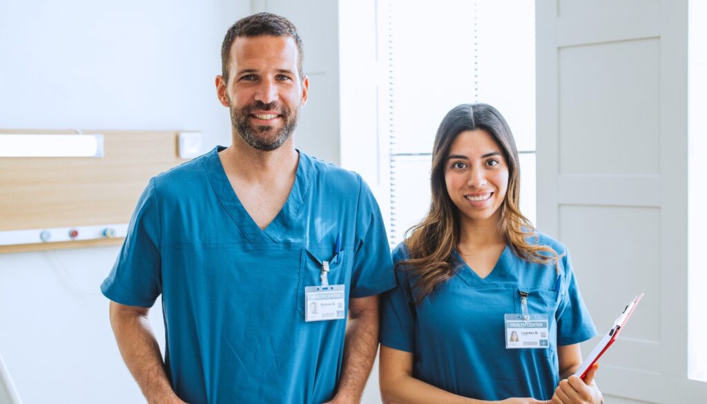 Two healthcare professionals in blue scrubs and ID badges stand indoors, smiling at the camera; one holds a clipboard.