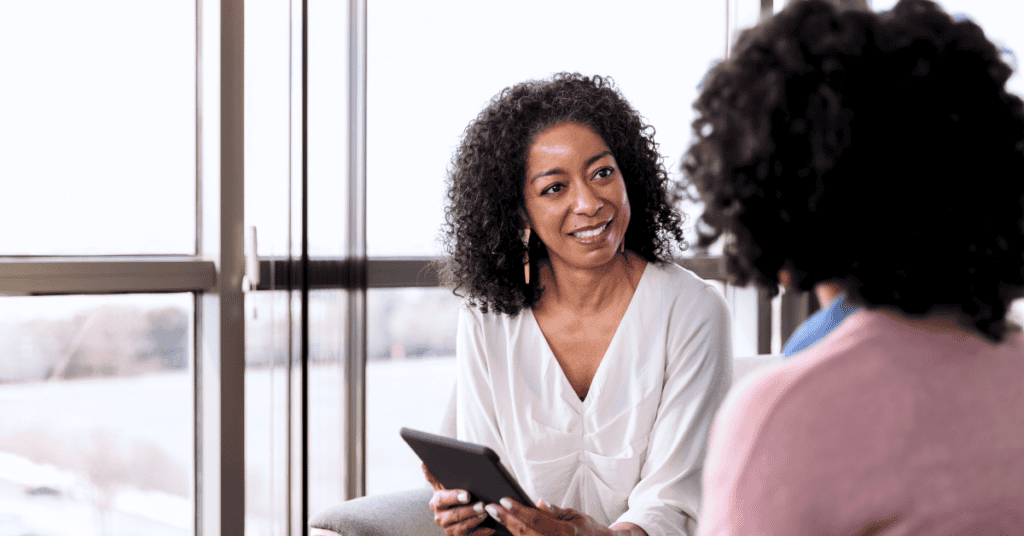 Two women sit indoors by large windows, one holding a tablet and smiling while listening to the other.