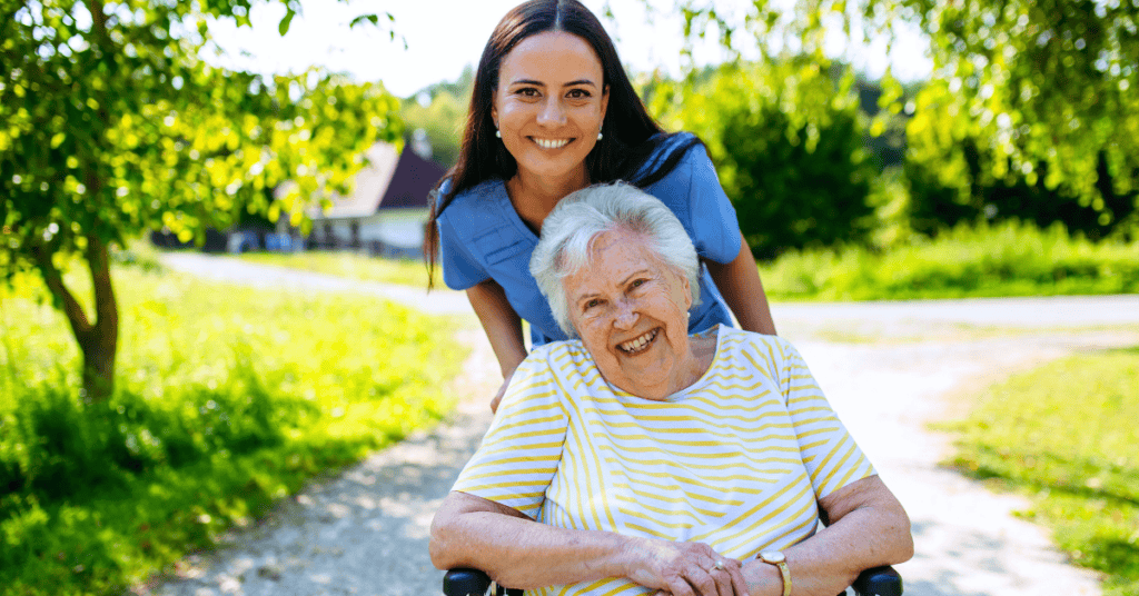 A younger woman stands behind an older woman in a wheelchair. Both are smiling outdoors on a sunny day with greenery in the background.