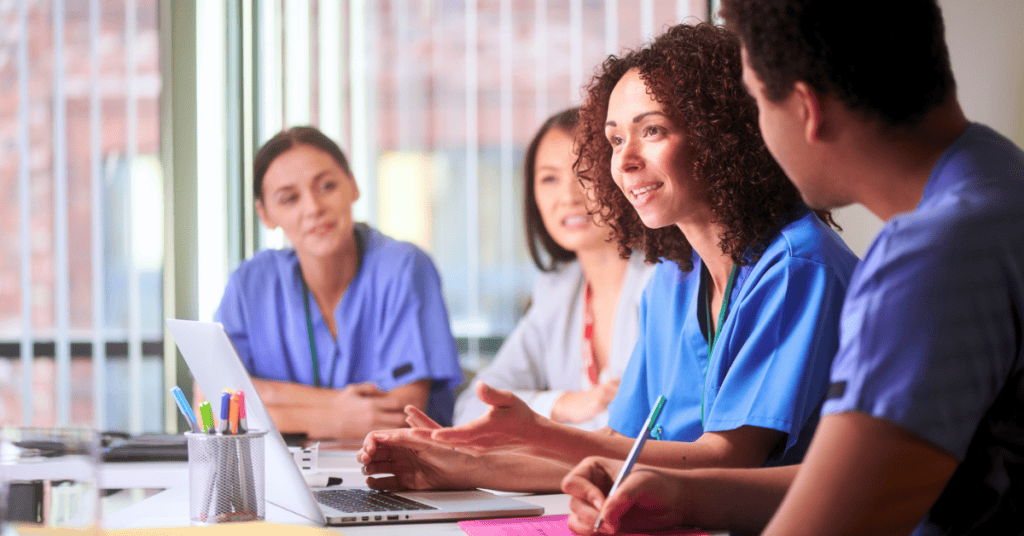 Four healthcare professionals in scrubs sit at a table with a laptop and papers, engaged in a discussion in a brightly lit meeting room.