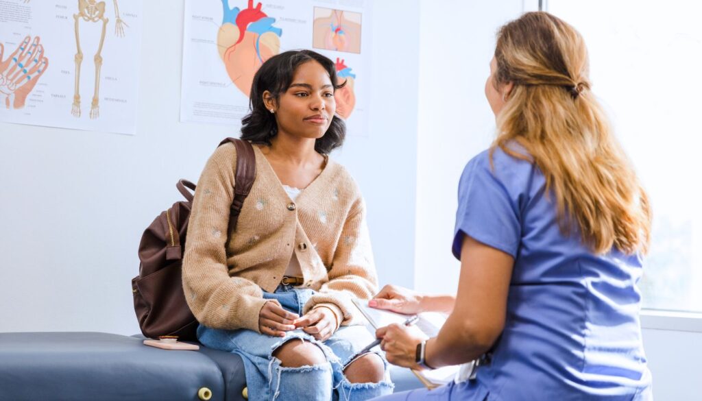 A young woman sits on an exam table with a backpack, speaking to a healthcare professional in scrubs in a medical office.