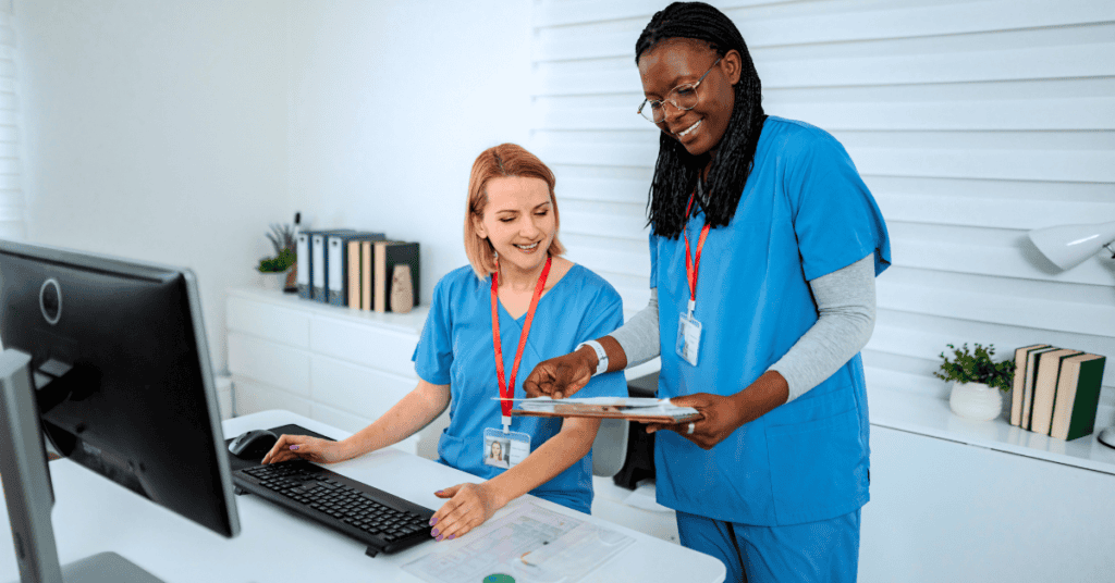 Two healthcare professionals in blue scrubs review documents together at a desk with a computer in a well-lit office setting.