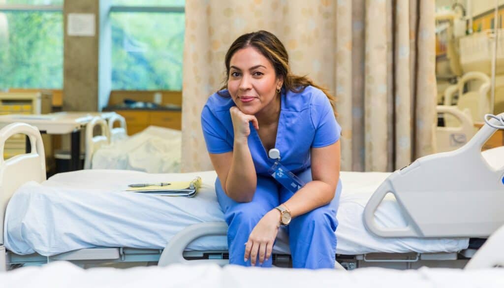 A nurse in blue scrubs sits on a hospital bed, resting her chin on her hand, with a clipboard nearby in a hospital room.