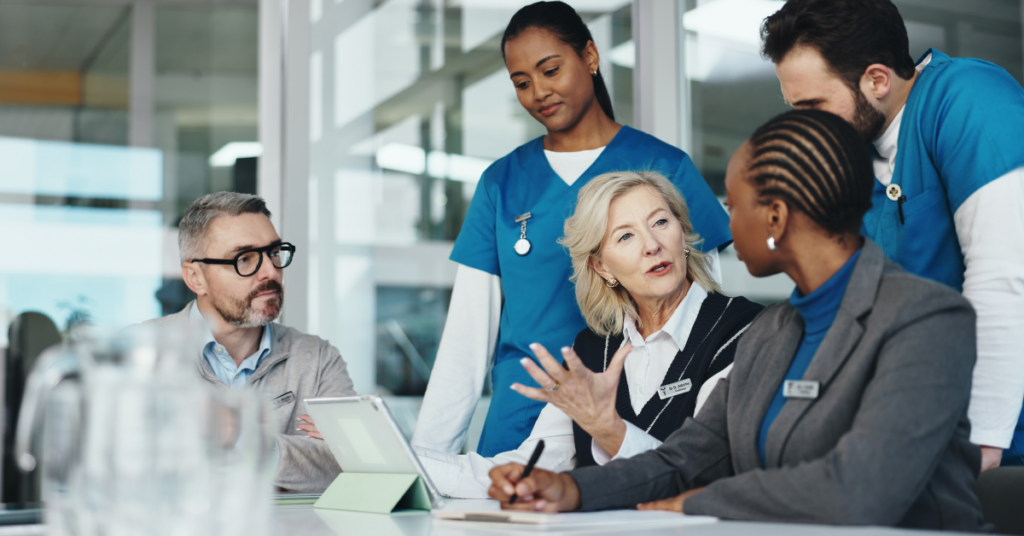 A group of healthcare professionals in discussion around a table, with two people in business attire and three in scrubs, in a modern office setting.