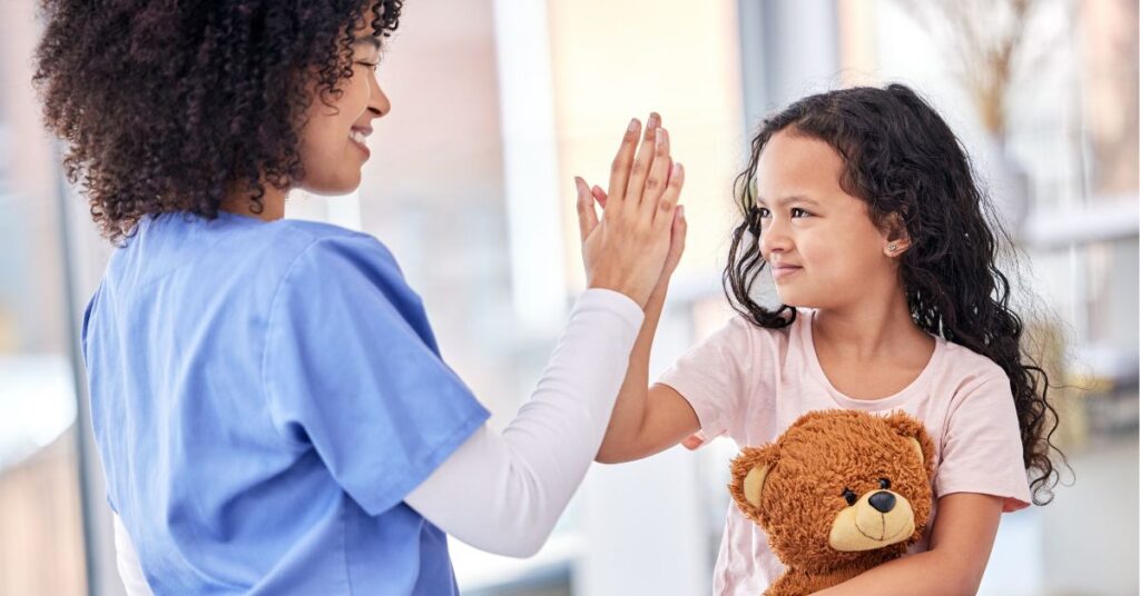 A nurse in blue scrubs gives a high-five to a young girl holding a teddy bear.
