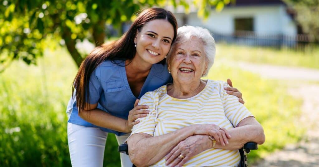 A young woman standing behind an older woman in a wheelchair, both smiling outdoors on a sunny day.
