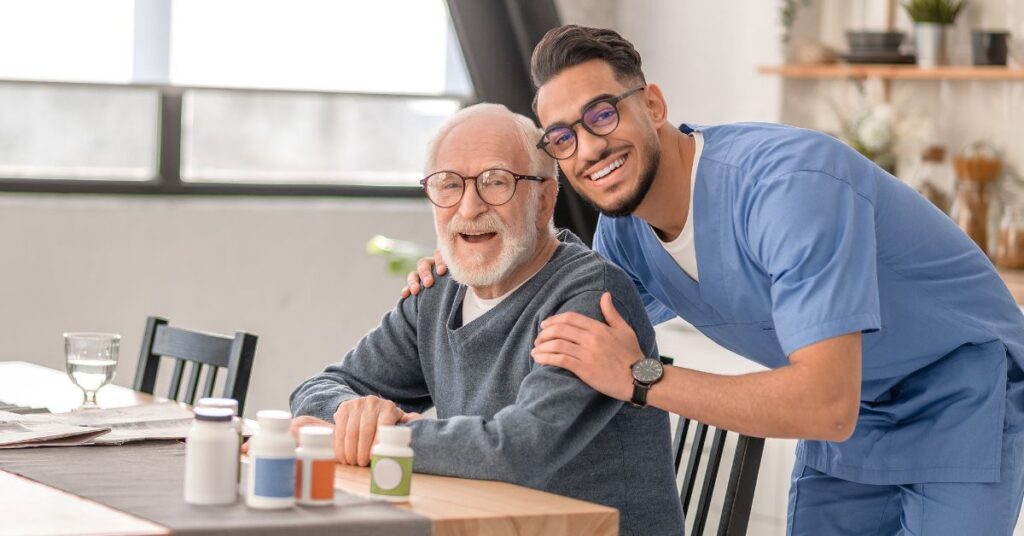 A smiling elderly man sits at a table with medication bottles while a young male caregiver in scrubs stands beside him, both smiling at the camera.