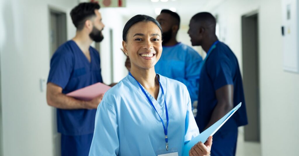 A woman in medical scrubs holds a folder and smiles at the camera, standing in a hospital hallway with three colleagues talking in the background.