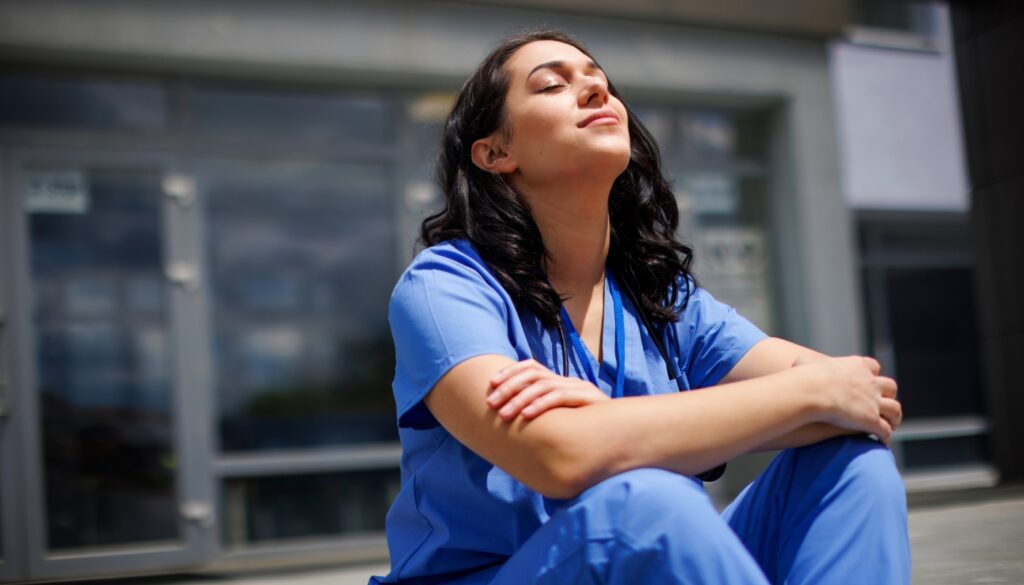 A person in blue medical scrubs sits outside with eyes closed and arms resting on knees, appearing relaxed in front of a building.