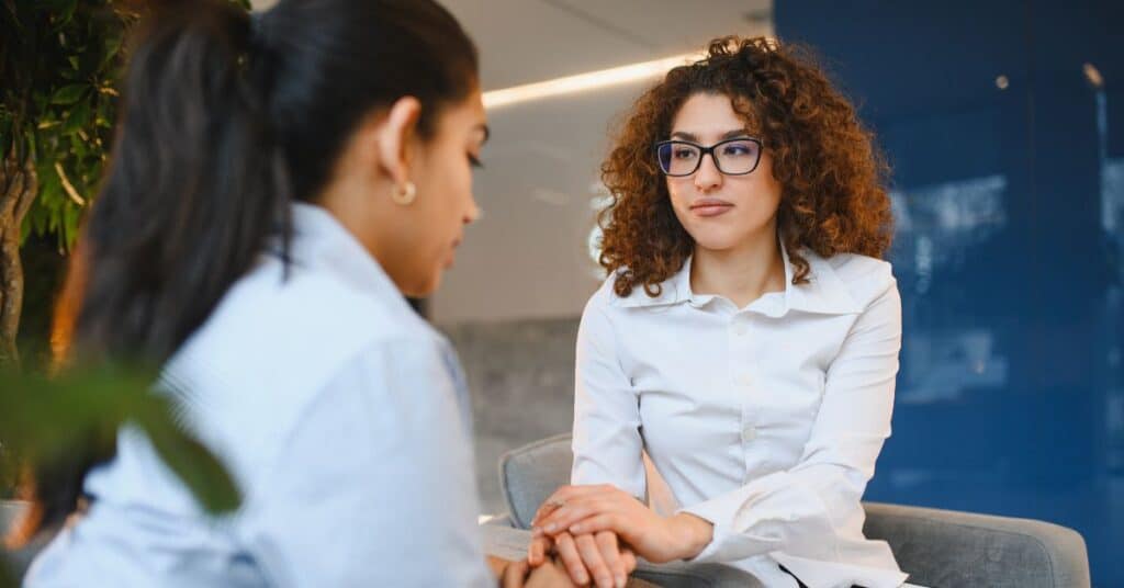 Two women sit facing each other indoors; one appears to be offering support by holding the other's hand while they talk.