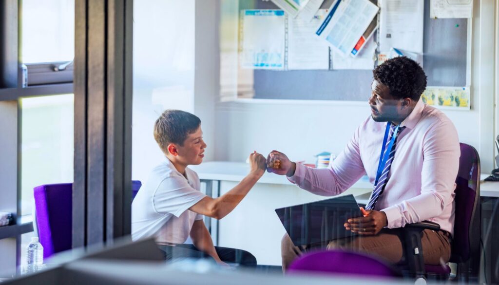 A man in a dress shirt and tie fist bumps a boy in a t-shirt while sitting together in an office or classroom setting.