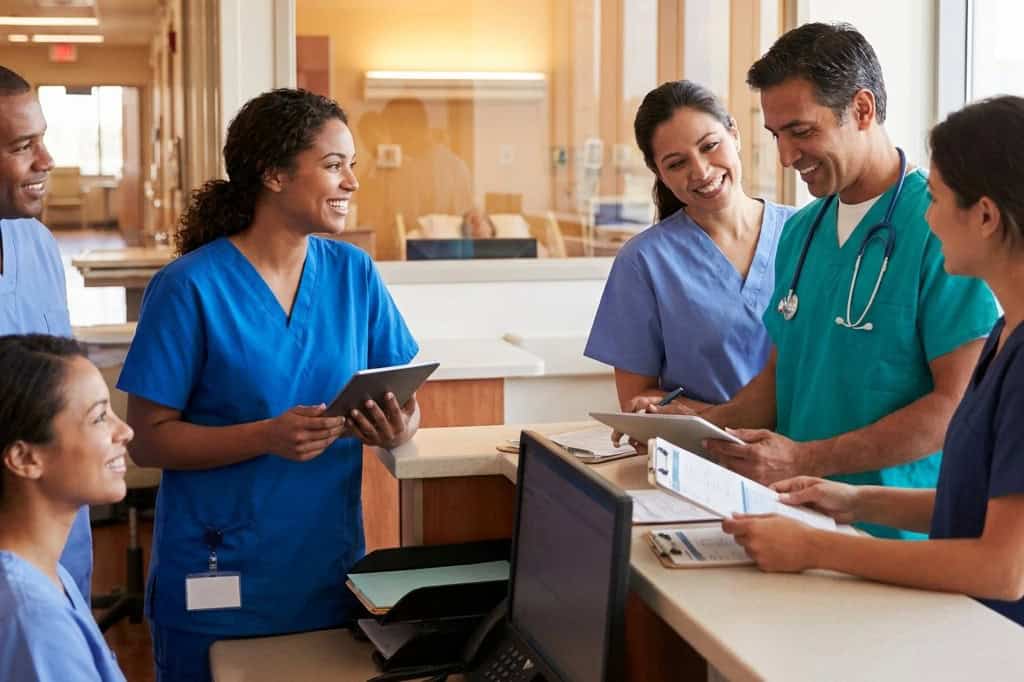 A group of medical professionals in scrubs stand around a hospital desk, discussing documents and using digital tablets.