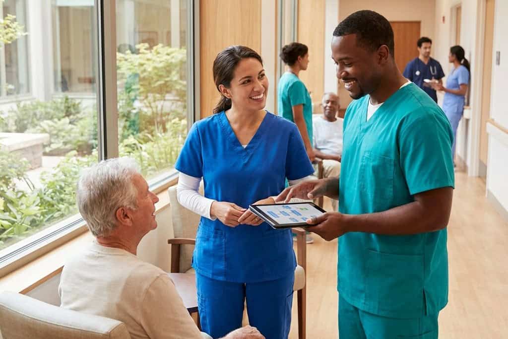 Two healthcare professionals review information on a tablet with an older male patient in a medical facility while others interact in the background.