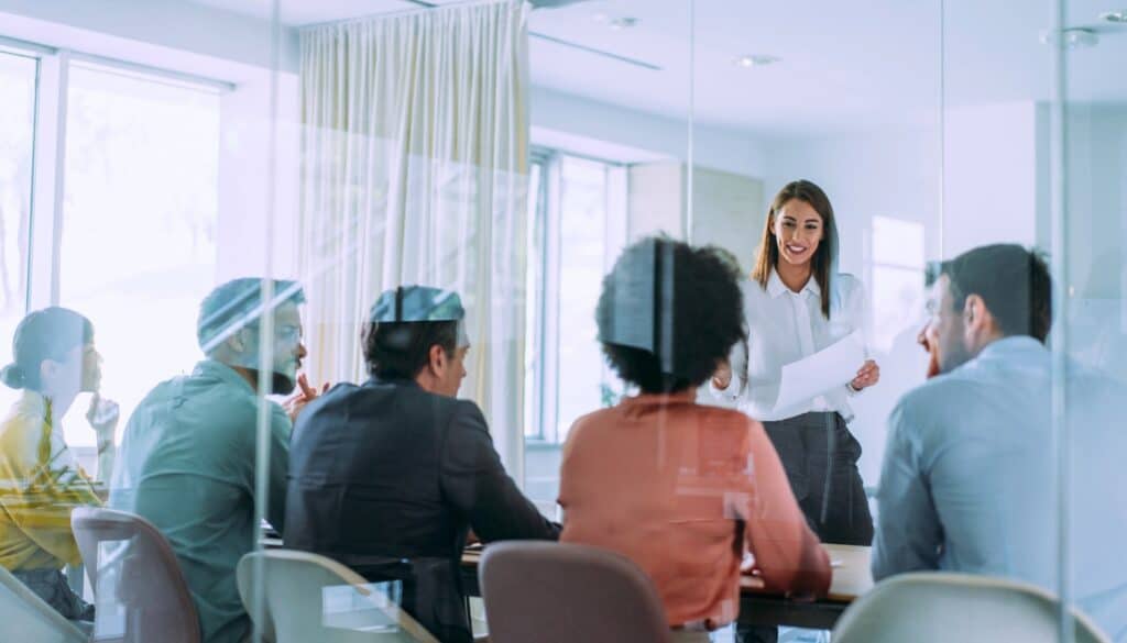 A woman stands and presents papers to a group of people seated around a conference table in a modern office meeting room.