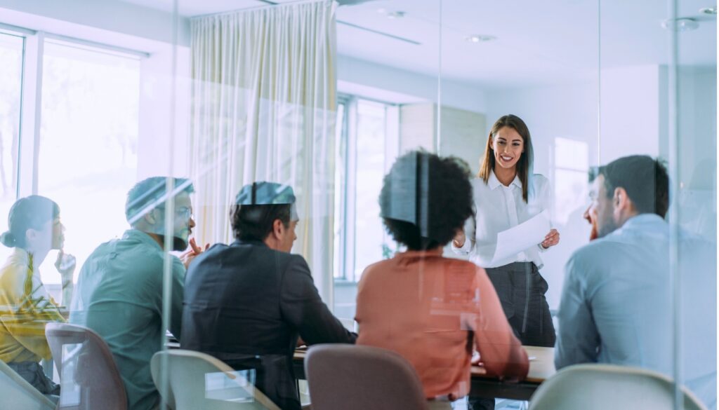 A woman stands and presents documents to a group of five people seated around a conference table in a modern office meeting room.