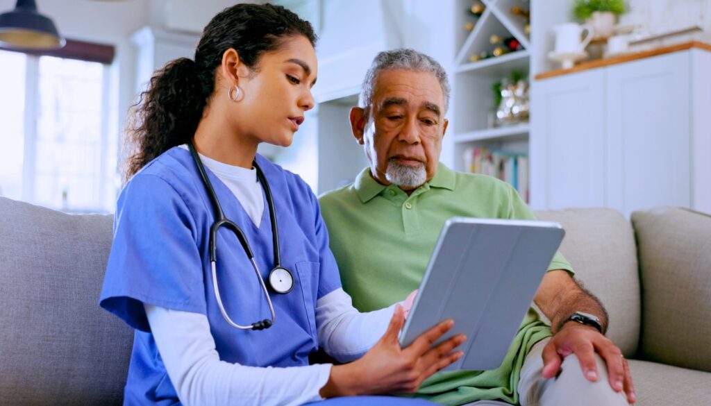 A healthcare professional in scrubs shows an older man information on a tablet as they sit together on a couch in a home setting.