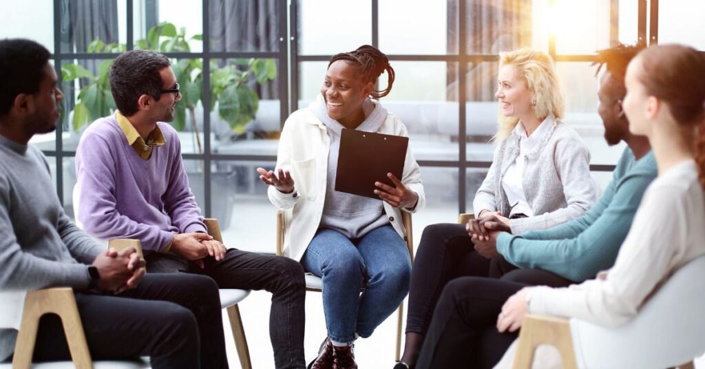 Six people sit in a circle in a bright room, engaged in a group discussion; one person holds a clipboard and speaks while others listen.