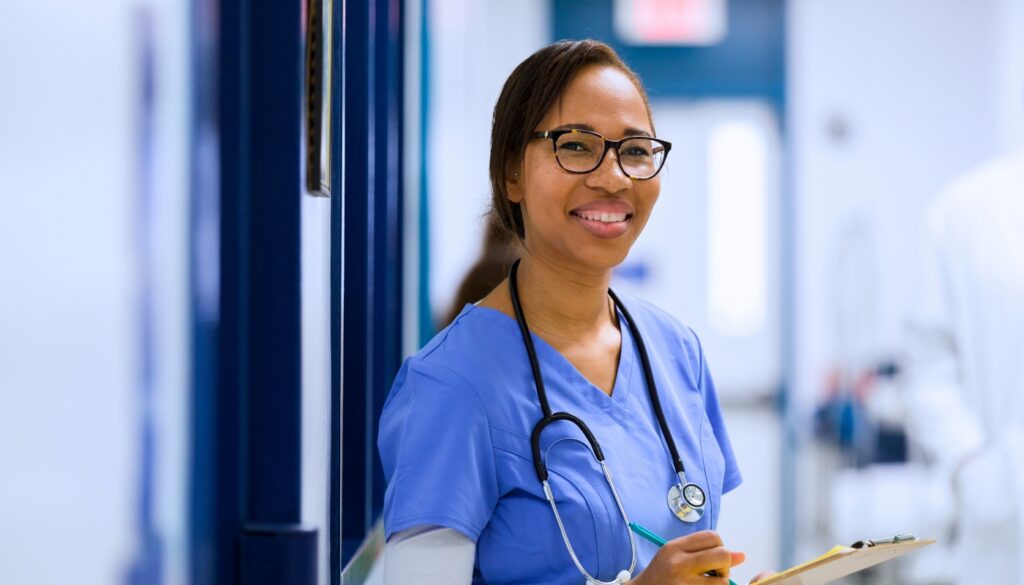 A healthcare worker in blue scrubs and a stethoscope stands in a hallway, holding a clipboard and smiling at the camera.