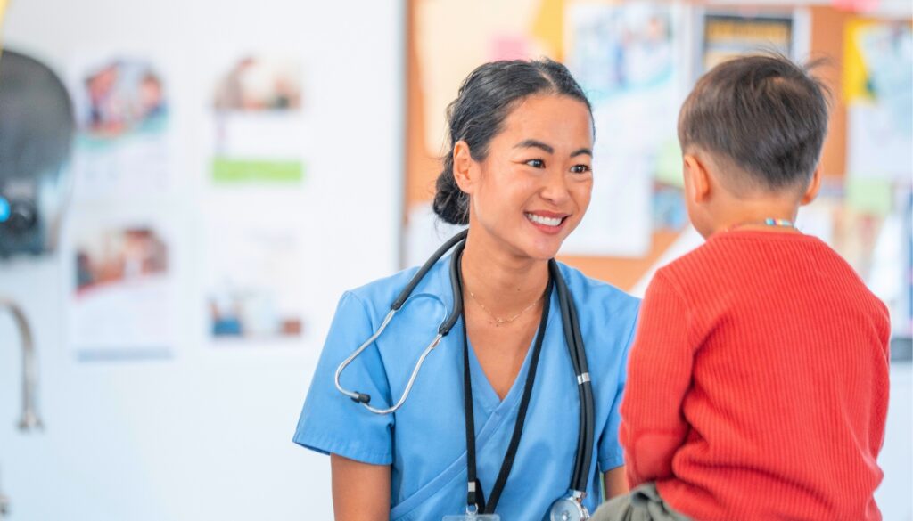 A healthcare worker in blue scrubs smiles while talking to a young child in a red sweater in a clinical setting.