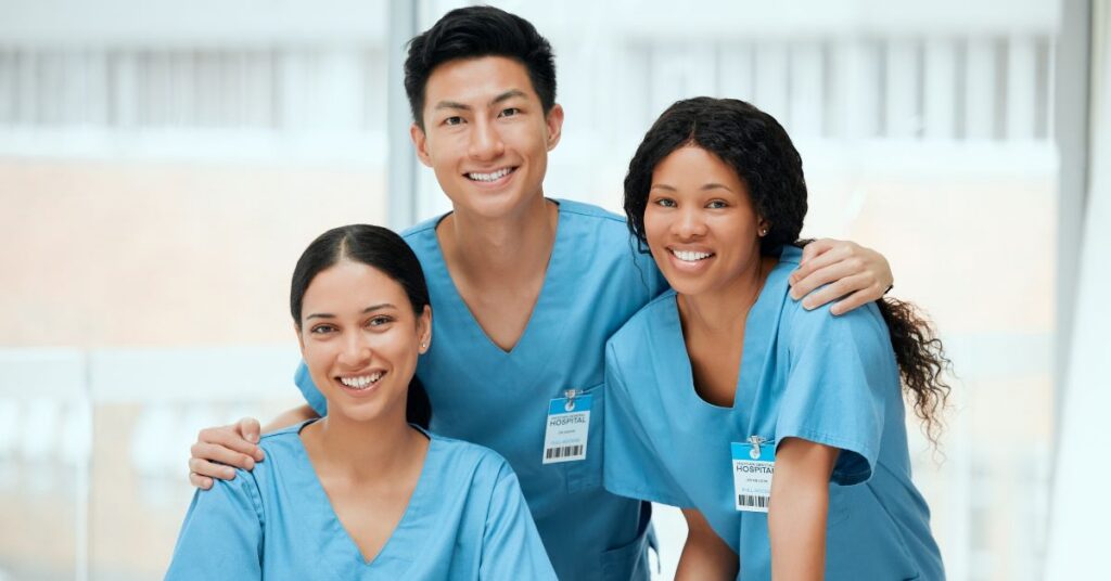 Three healthcare professionals in blue scrubs and hospital ID badges pose together, smiling at the camera in a bright indoor setting.