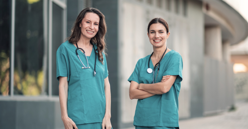 Two women in teal medical scrubs, each with a stethoscope, standing outside a modern building and smiling at the camera.