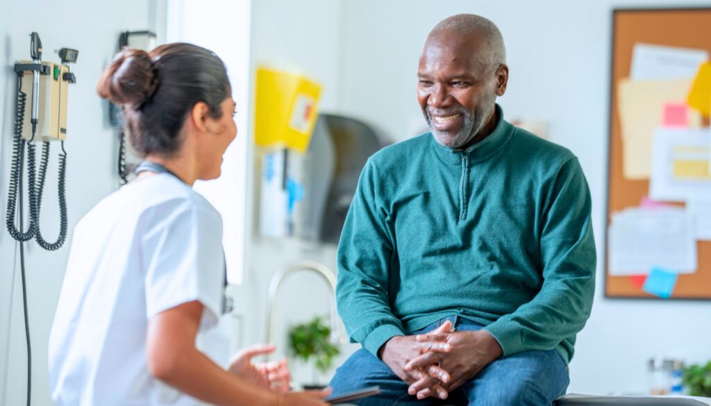 A doctor talks with a smiling man who is sitting on an exam table in a medical office.
