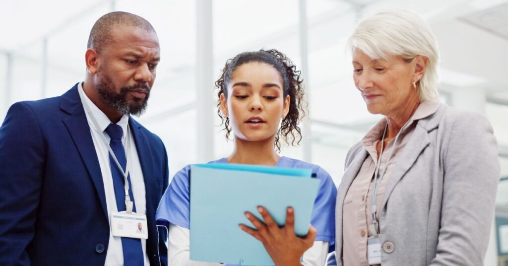 Three professionals, including a nurse and two colleagues with badges, stand together in an office, reviewing documents on a clipboard.
