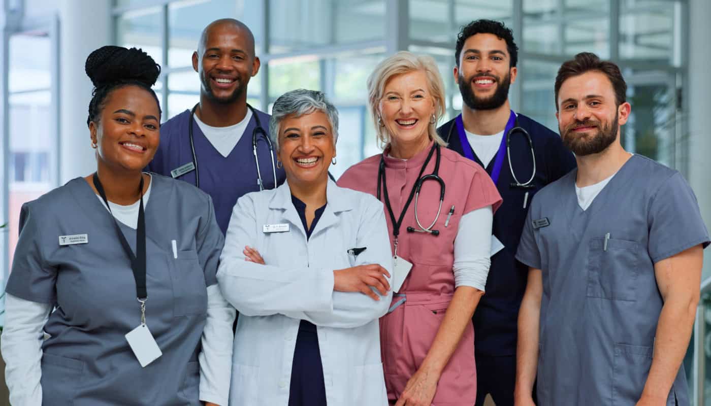 Six medical professionals in scrubs and lab coats stand together indoors, smiling at the camera, with stethoscopes and ID badges visible.