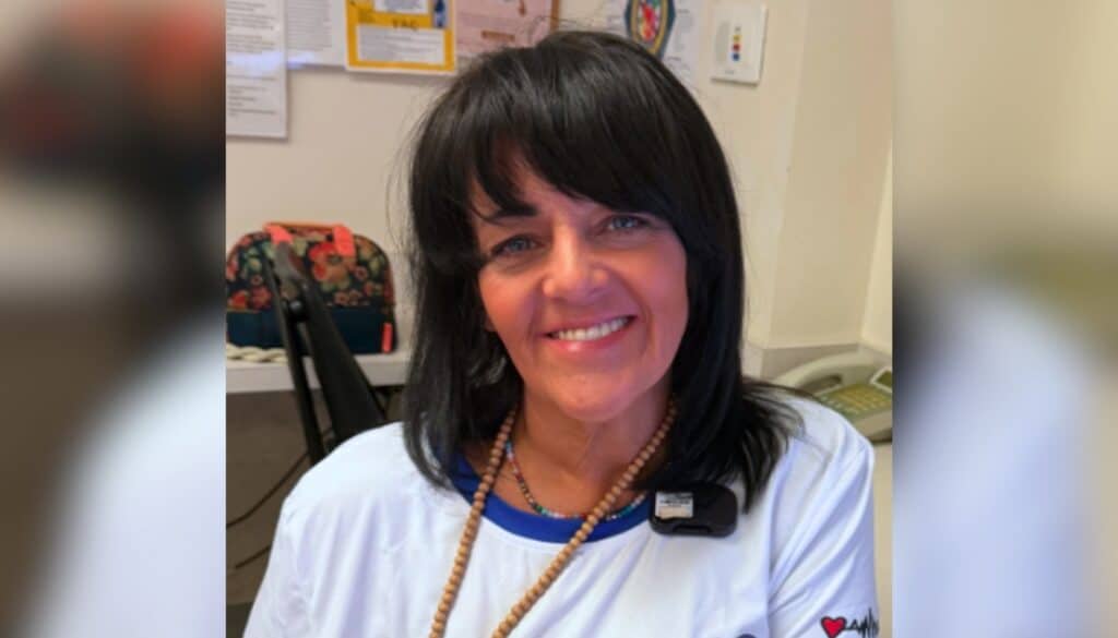 Woman with dark hair and a white shirt sits indoors, smiling at the camera; office items and a floral bag are visible in the background.