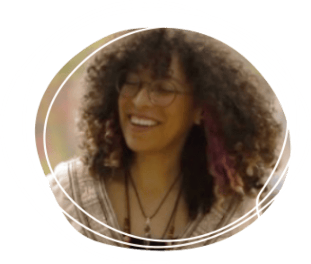 Smiling person with curly hair, glasses, and layered necklaces, wearing a patterned top, photographed outdoors with a circular white graphic overlay.
