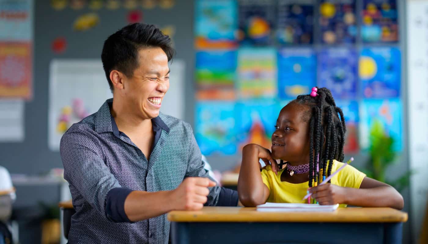 A man and a young girl sit at a desk in a colorful classroom, smiling at each other. The girl holds a pencil, and papers are in front of them. Artworks hang on the wall in the background.