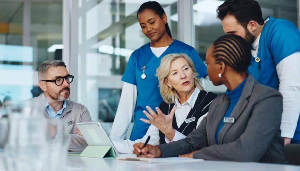 Five healthcare professionals, including nurses and administrators, sit and stand around a table in discussion, with paperwork, a tablet, and notepads visible.