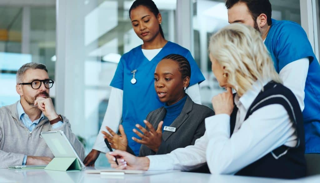 Five professionals, including two in medical scrubs and three in business attire, sit around a table having a discussion, with a tablet visible in front of them.