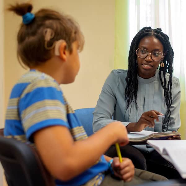A woman with glasses talks to a child wearing a hearing aid. The child holds a pencil, and they appear to be in a classroom or office setting.