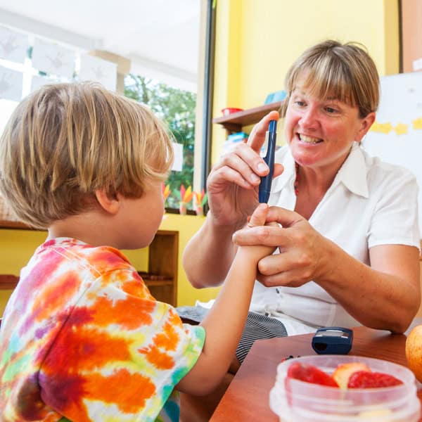 An adult checks a child’s blood sugar with a glucose meter in a classroom setting. Fruit is visible on the table.
