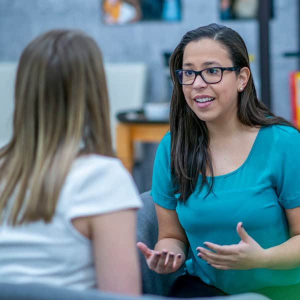 Two women sit facing each other indoors, engaged in a conversation. One woman talks with expressive hand gestures while the other listens.