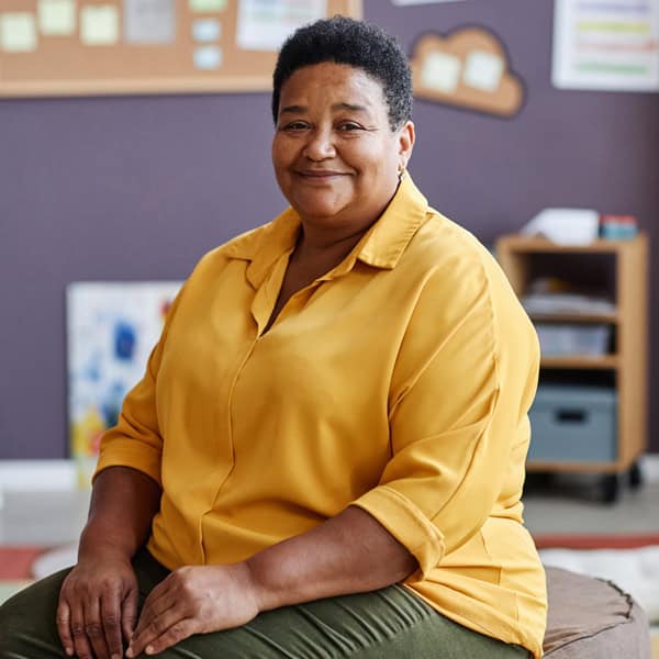 A person with short hair wearing a yellow shirt sits indoors, smiling at the camera. The background features bulletin boards and shelves with classroom materials.