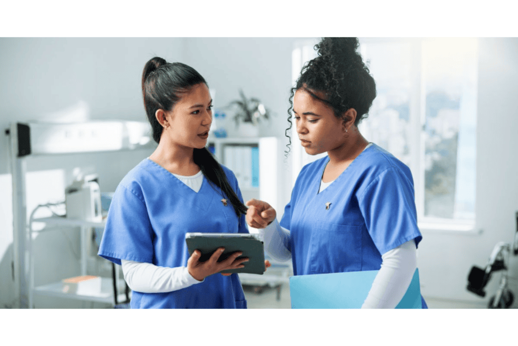 Two healthcare workers in blue scrubs discuss information on a tablet in a medical office setting, one holding a file folder.