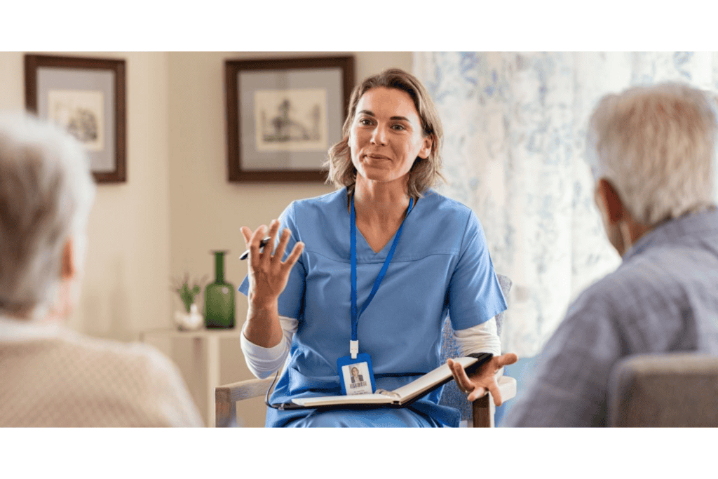 A healthcare professional in blue scrubs speaks with two older adults, holding a notebook and pen in a well-lit room.