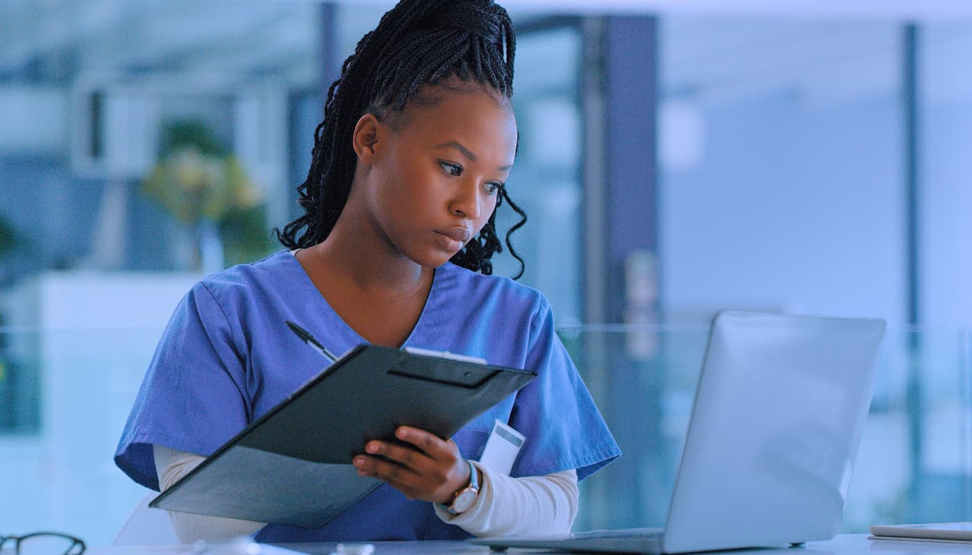 A nurse in blue scrubs reviews a clipboard while looking at a laptop in a clinical setting.