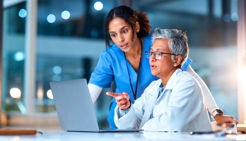 Two healthcare professionals look at a laptop screen together in a bright, modern office setting. One is seated and pointing at the screen, while the other stands and listens attentively.