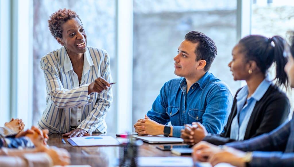 A woman stands and talks to colleagues seated around a table in a modern office meeting room.